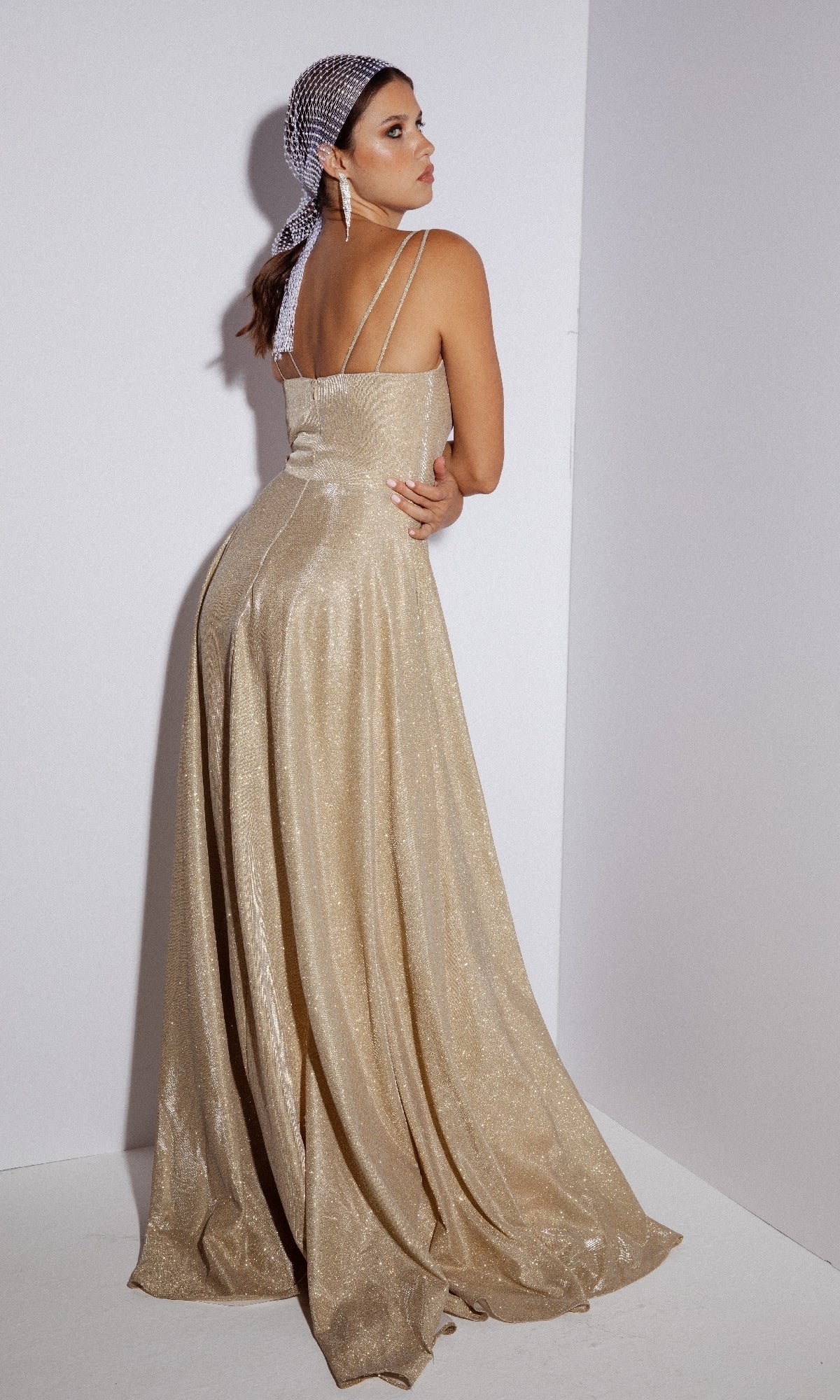 A woman stands in a corner facing away, wearing the Eureka 8900 Sleeveless Long Glitter Formal Dress in gold with thin straps and a silver headscarf.